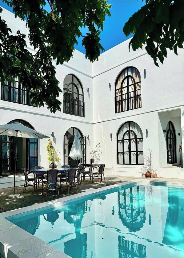 Un patio con piscina, mobiliario exterior y paredes blancas con ventanales en arco.