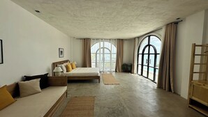 A spacious bedroom with a large bed, a sofa, and a wooden chair. There are two large windows with curtains, and a view of the outdoors is visible. - Uli Cliffhouse (Dasol)