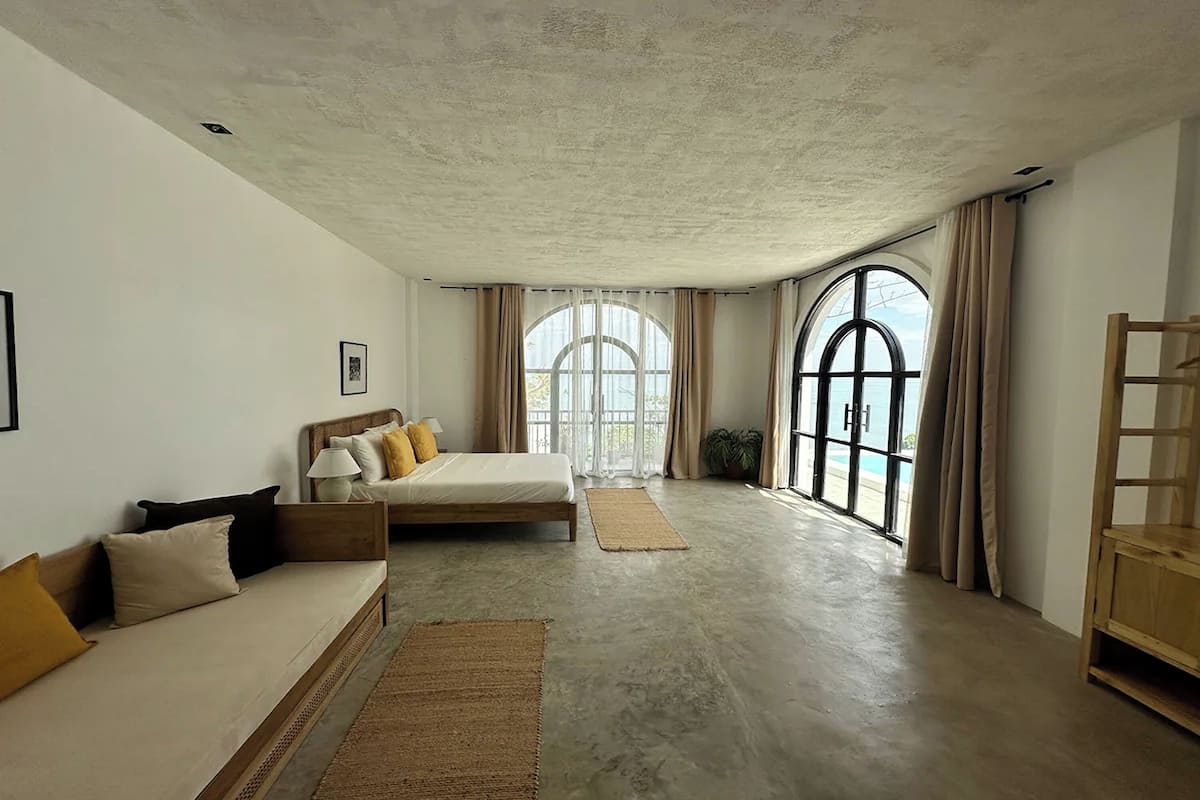 A spacious bedroom with a large bed, a sofa, and a wooden chair. There are two large windows with curtains, and a view of the outdoors is visible.