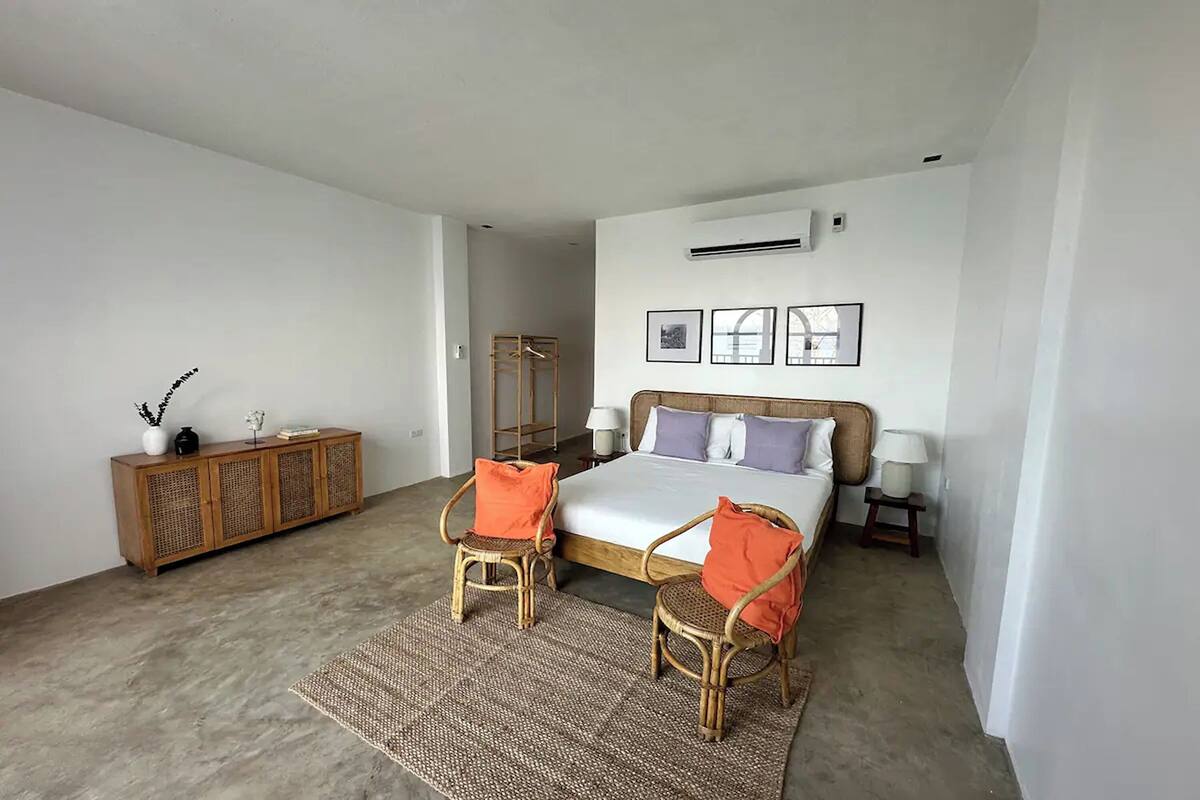 A bedroom with a bed, two wicker chairs, a wooden cabinet, and a small table with a lamp.
