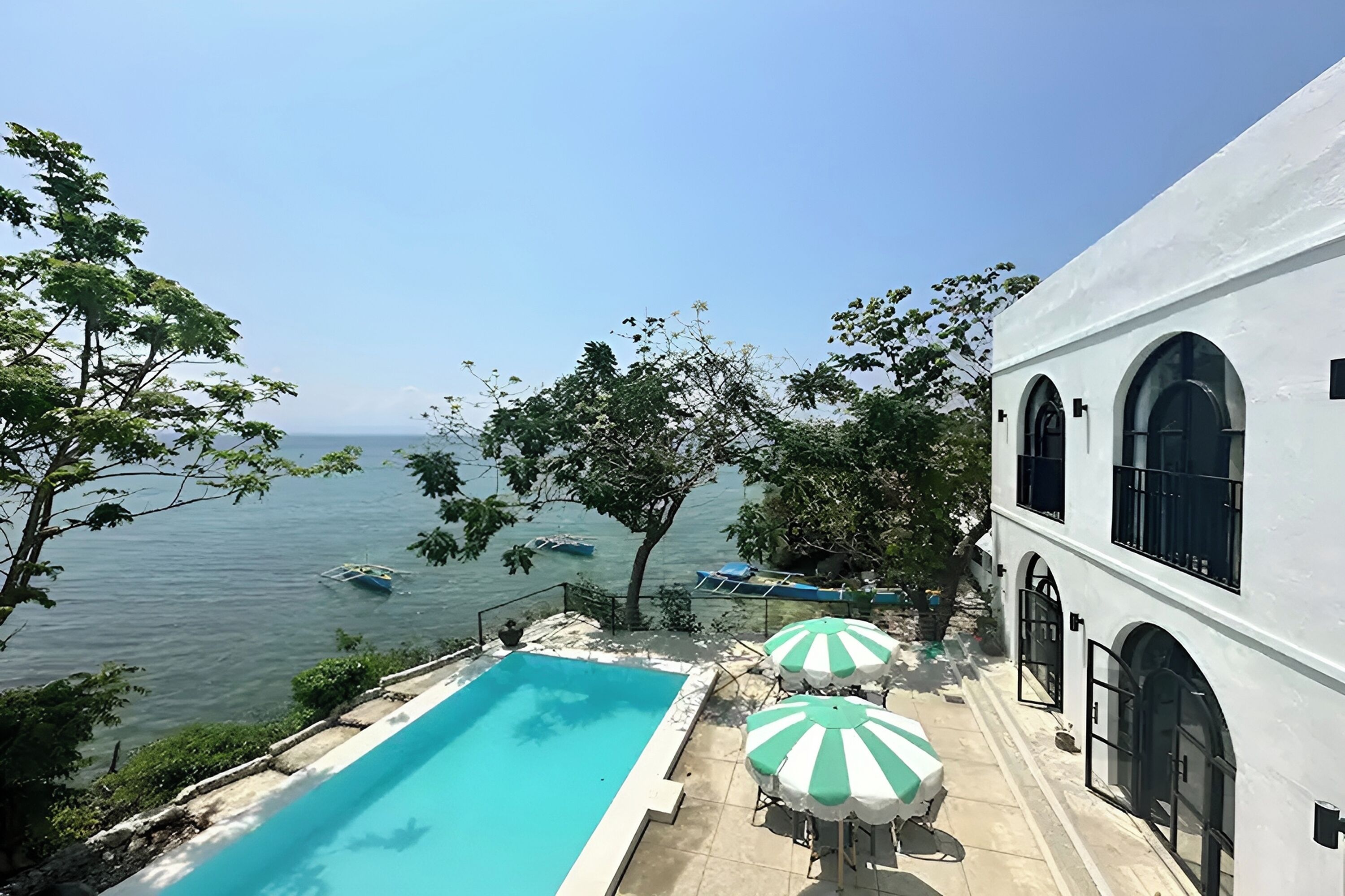 A pool area with a view of the sea, two umbrellas, and a white building with arched windows.