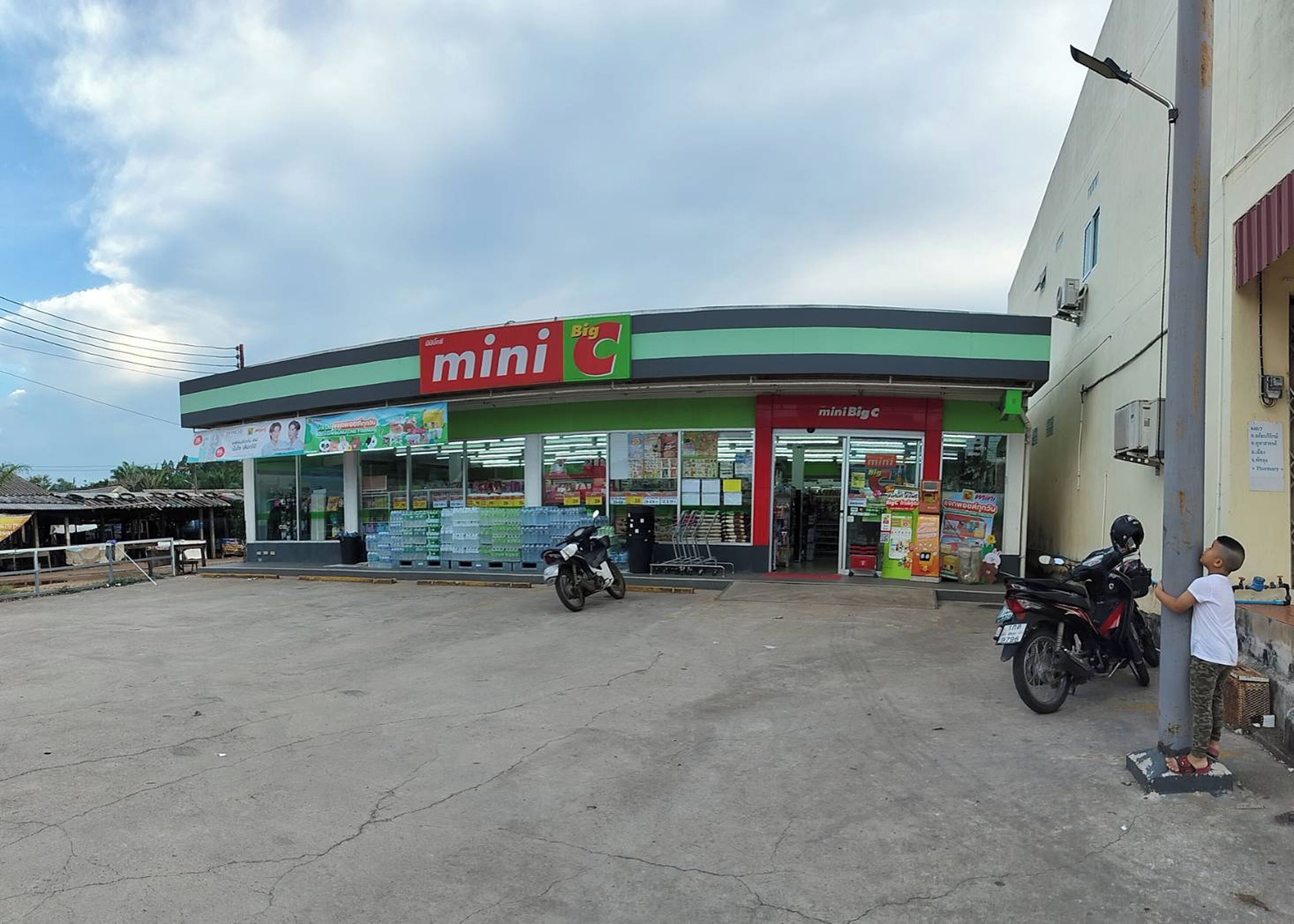 A mini Big C convenience store with a clear entrance, a motorcycle parked in front, and a child standing by the entrance.