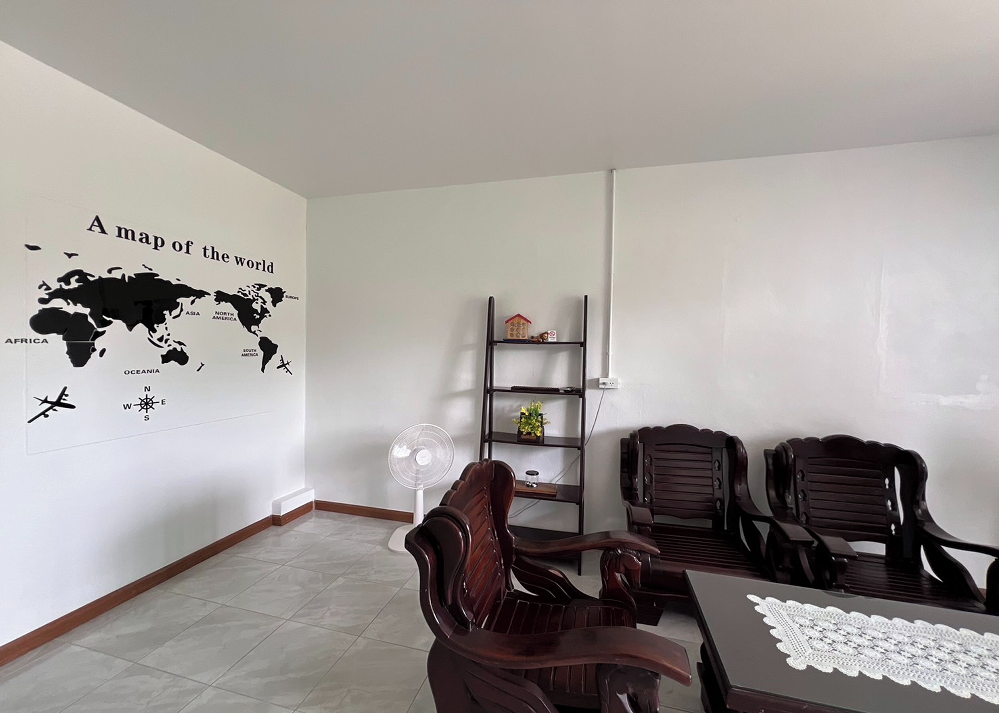 Un espacio de oficina moderno con un mapa mundial en la pared, un escritorio con una computadora portátil, dos sillas de madera, un estante pequeño con adornos y un ventilador de pie.