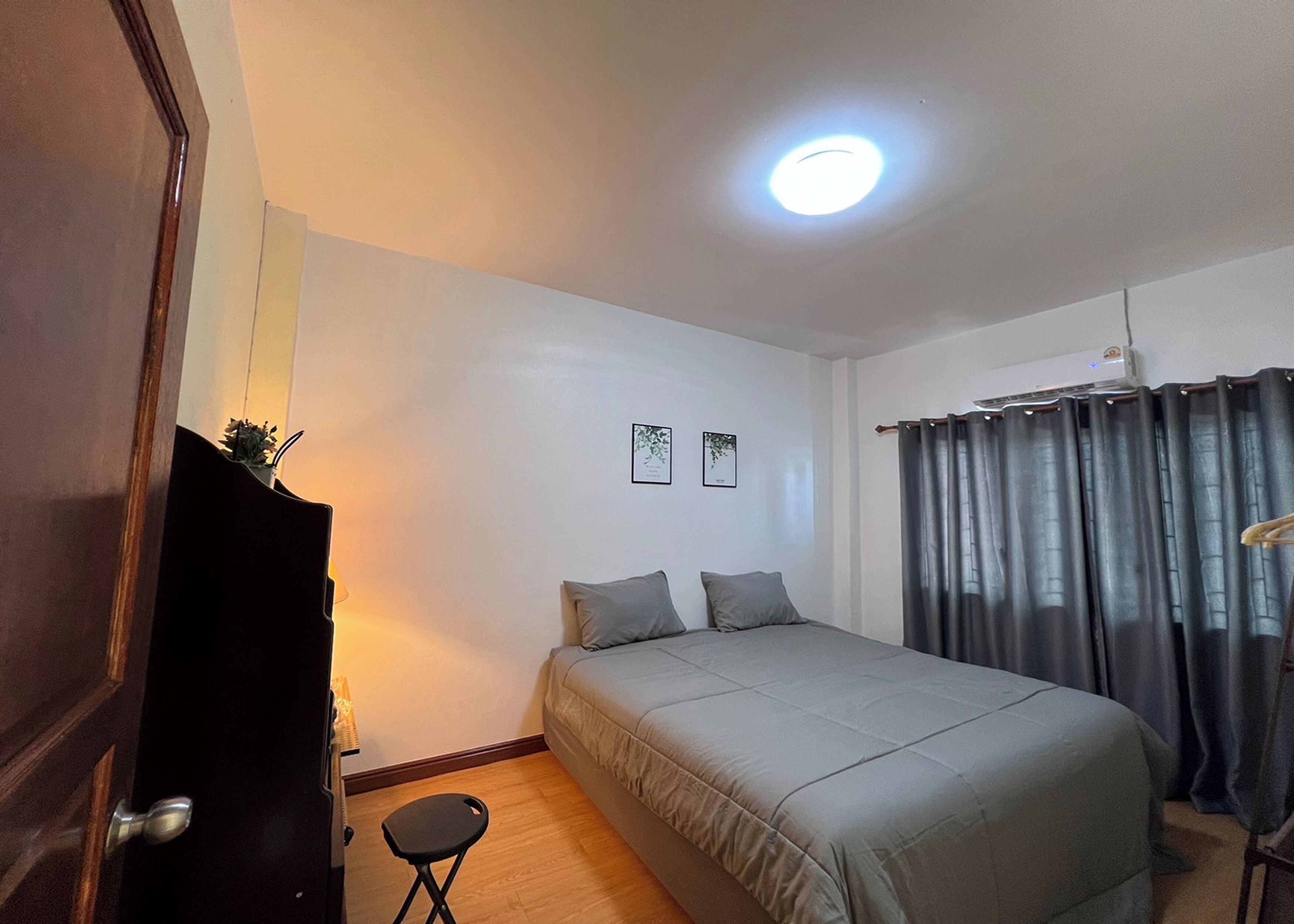 A bedroom with a bed, a small stool, a dark door, a ceiling light, and a window with curtains.