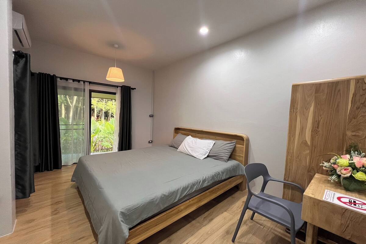 Quarto individual com uma cama e cabeceira de madeira, edredom cinza, uma cadeira preta, uma mesa de madeira com um vaso de flores, e vista para o verde através de uma porta deslizante.