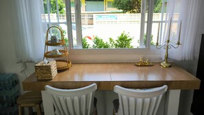Une pièce avec une table en bois, deux chaises blanches, une étagère en osier, un chandelier et une vue sur un bâtiment et de la verdure à l’extérieur.
