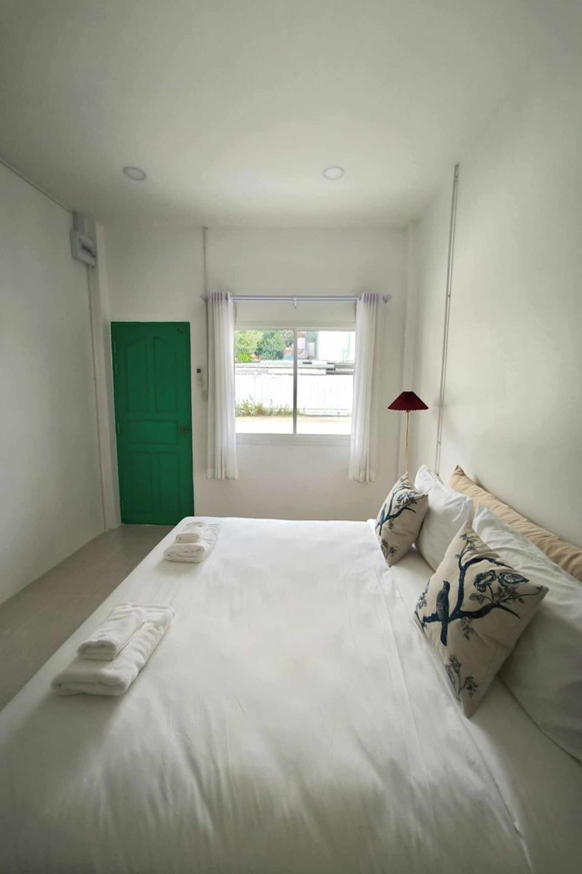 A hotel room with a bed, a green door, a window with curtains, and a red lamp.