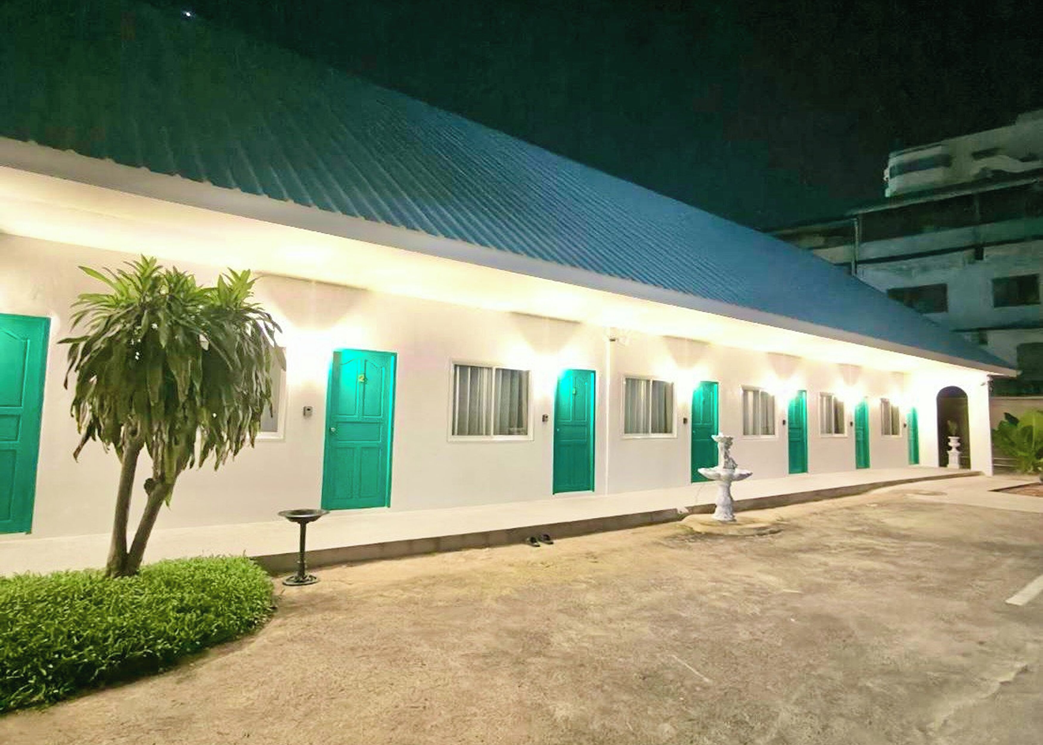A building with a row of rooms, each with a green door and windows, and a small garden area with a palm tree.