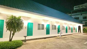 A building with a row of rooms, each with a green door and windows, and a small garden area with a palm tree.