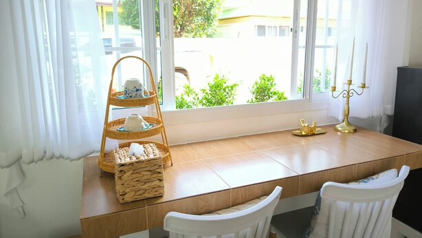 Une pièce avec une table en bois, deux chaises blanches, un plateau en osier contenant des tasses à thé, un chandelier et une vue sur la verdure et les bâtiments à l’extérieur.