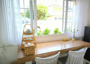 Ein Zimmer mit einem Holztisch, zwei weißen Stühlen, einer Korbtasse mit Teetassen, einem Kandelaber und Blick auf Grünflächen und Gebäude.