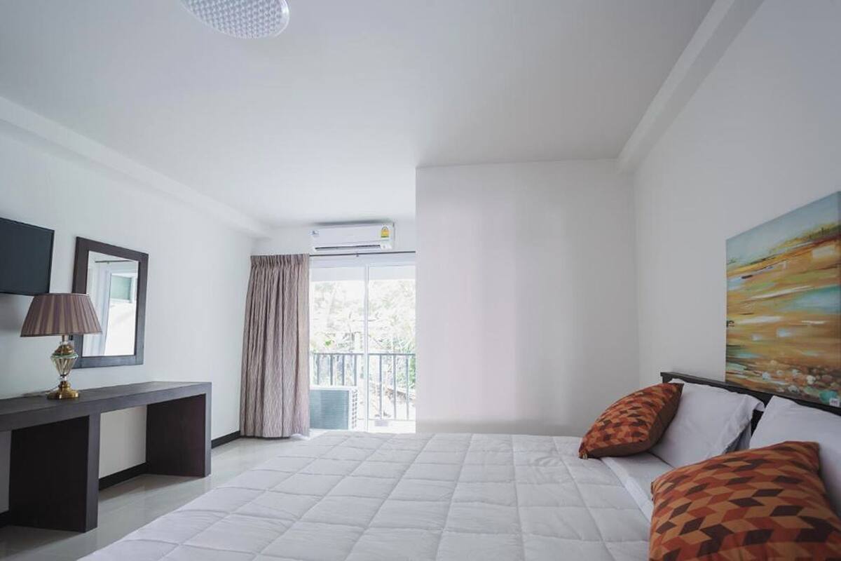 Un dormitorio con cama, escritorio, lámpara, un cuadro y vistas al exterior.