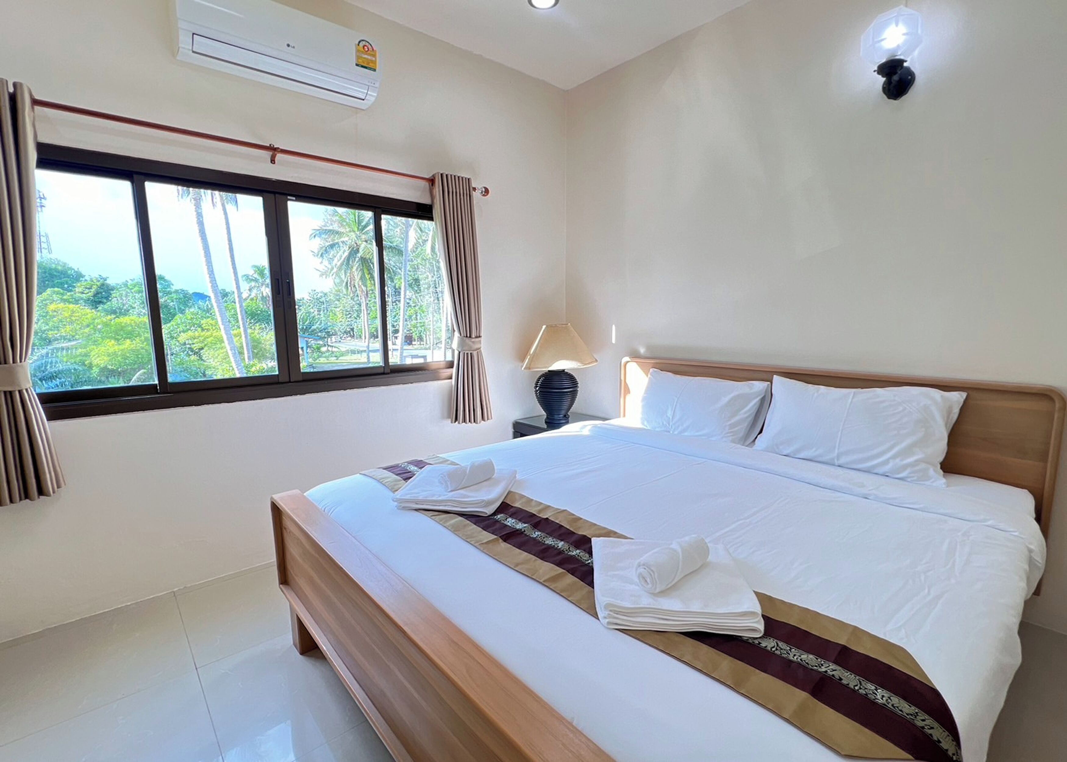 A hotel room with a large bed, a nightstand with a lamp, a window with curtains, and an air conditioning unit.