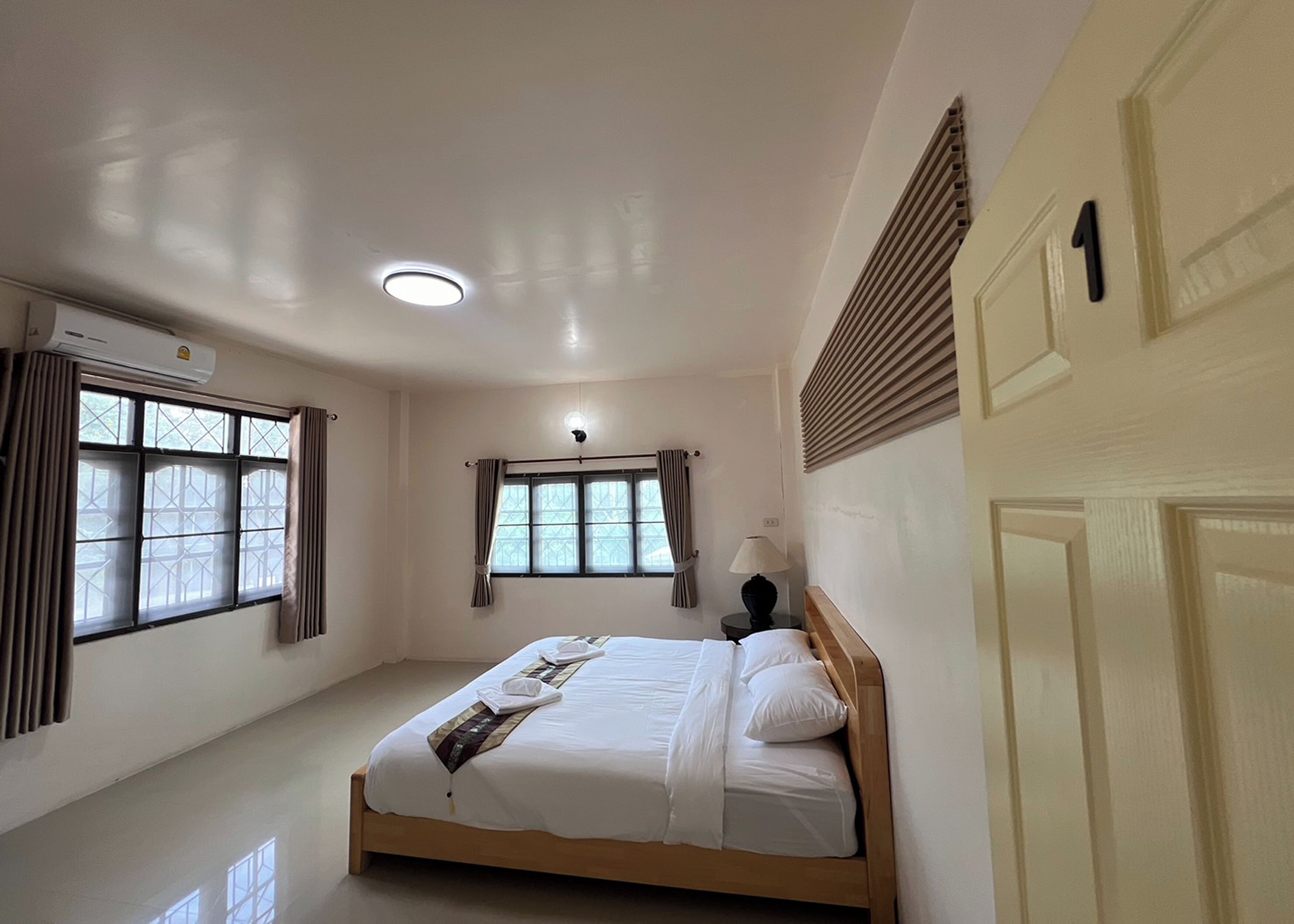 A bedroom with a bed, two windows with curtains, an air conditioning unit, and a ceiling light.
