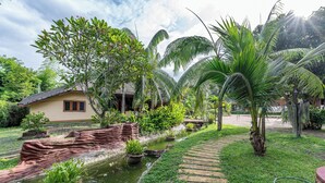 Un jardin tropical avec un bassin, des palmiers et une maison au toit recouvert de tuiles.