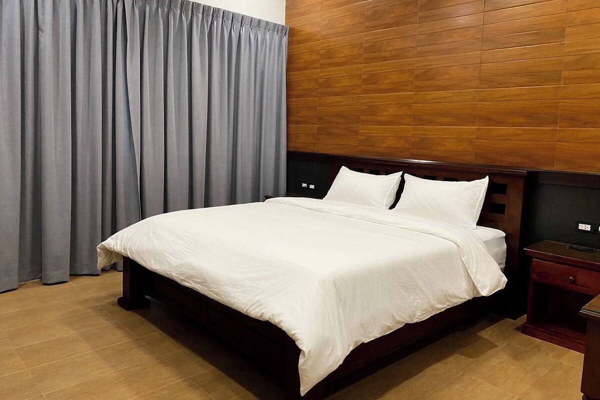 A neatly made bed with white linens in a room with wooden paneling on the walls, a wooden nightstand, and a grey curtain.