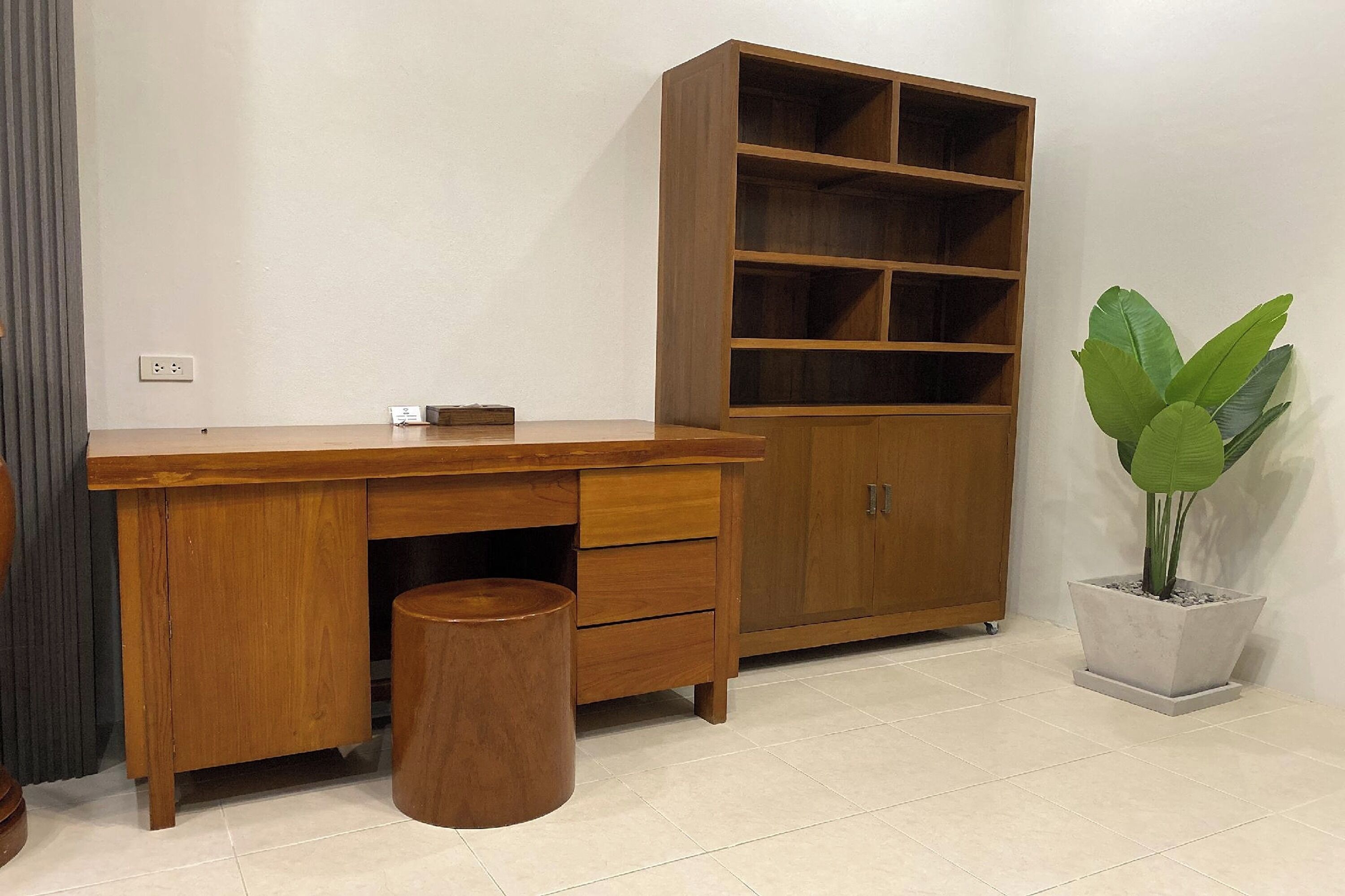 Een houten bureau met laden en een prullenbak, een groot houten kast met meerdere planken, en een potplant.