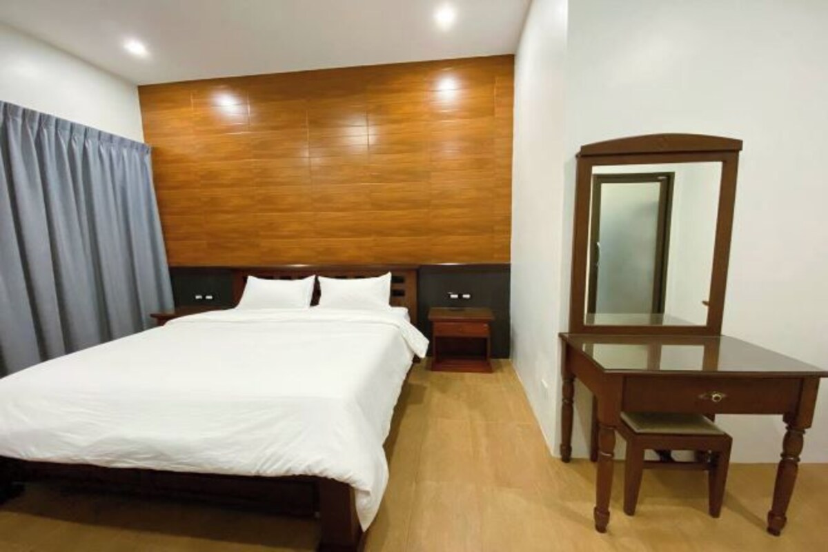 Une chambre d’hôtel avec une tête de lit en bois, un grand lit avec du linge de lit blanc, une table de chevet en bois, une commode en bois avec un miroir et une petite chaise en bois.