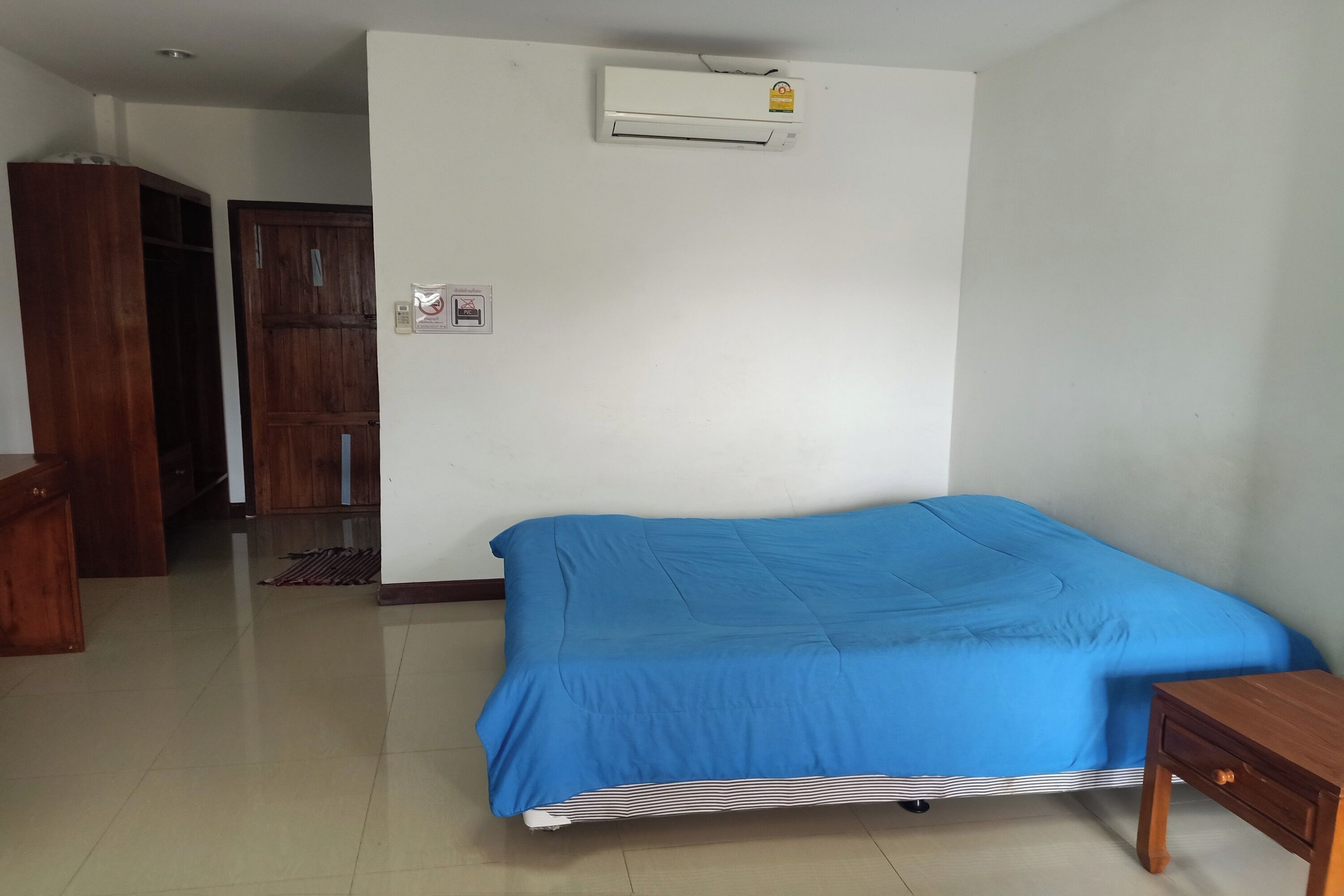 A bedroom with a bed, a wooden wardrobe, a door, and an air conditioning unit on the wall.