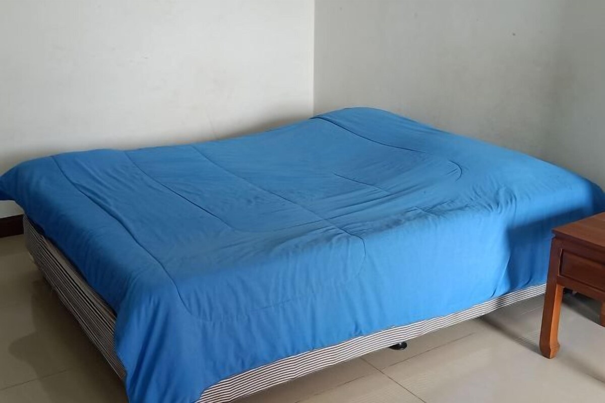 A single bed with a blue bedspread in a room with plain walls and a wooden nightstand.