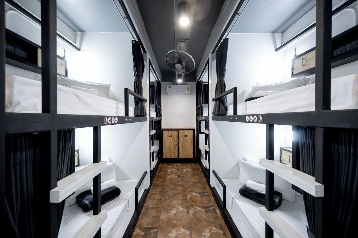 Un couloir étroit, avec des lits superposés de chaque côté, un appareil de climatisation central et une porte en bois au fond.