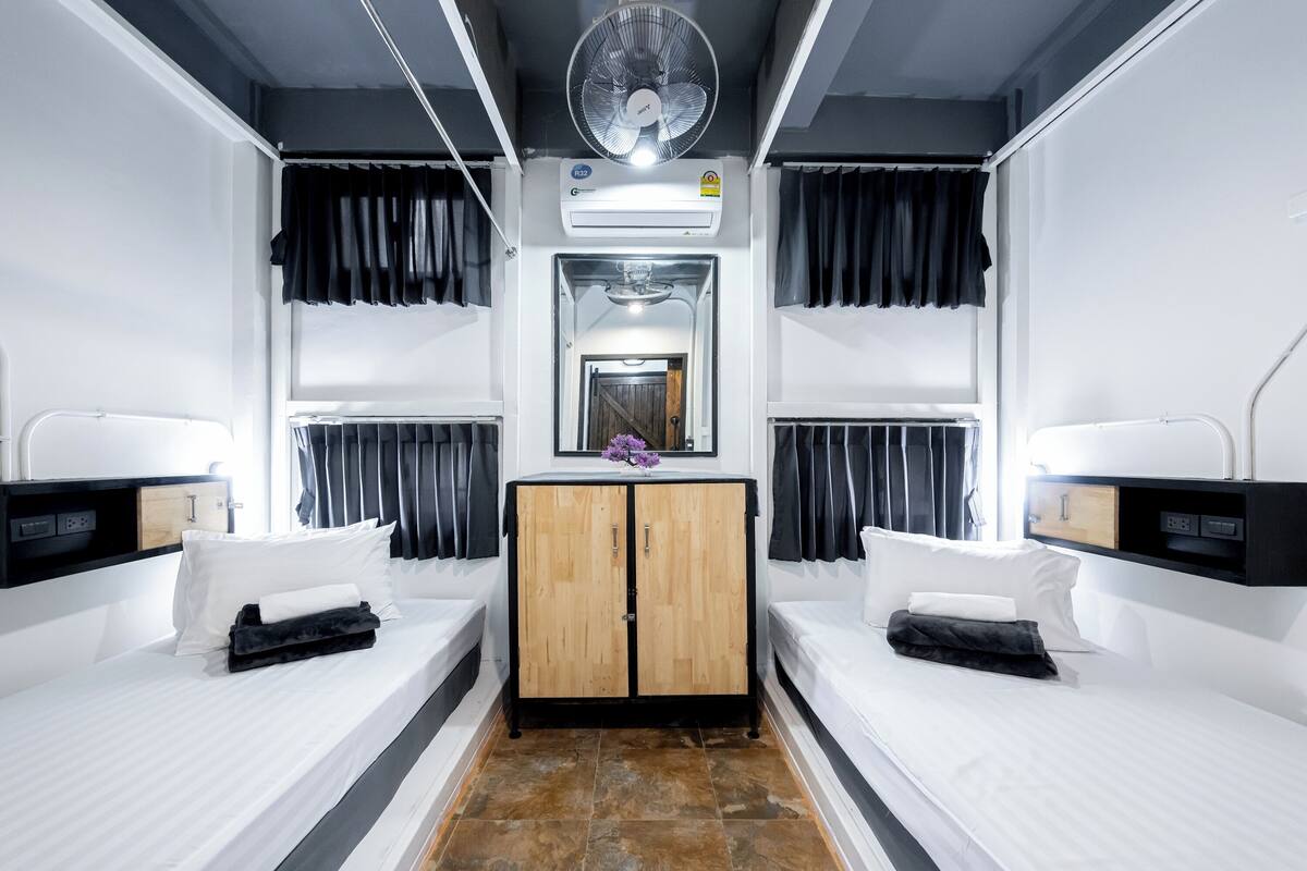 Une chambre d’hôtel avec deux lits, une armoire en bois, un miroir et un ventilateur de plafond.