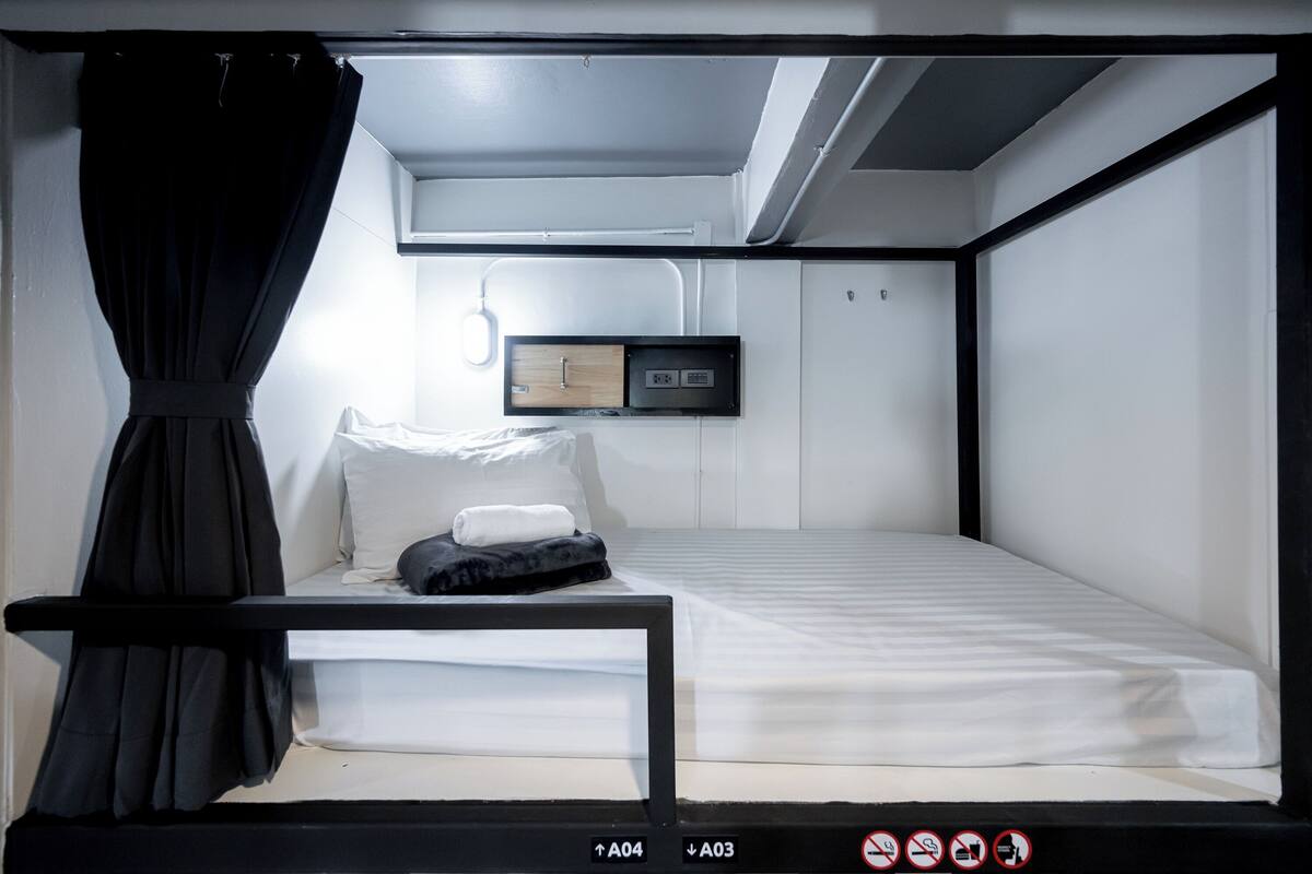 Un lit superposé avec une structure noire, une literie blanche et une petite table de chevet. Une prise de courant et un panneau de commande sont fixés au mur au-dessus du lit.