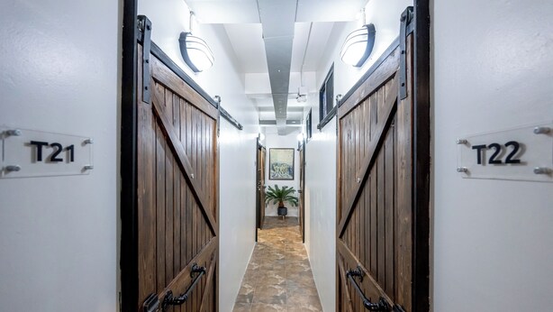 Un couloir avec des portes en bois portant les numéros T21 et T22, qui mène à une pièce au sol en pierre et où se trouve une plante en pot.