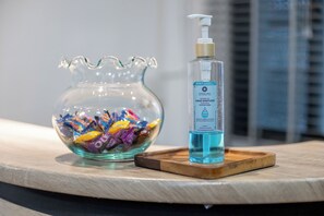 Un tazón de vidrio transparente lleno de caramelos de colores y una botella de gel antibacterial sobre una bandeja de madera.