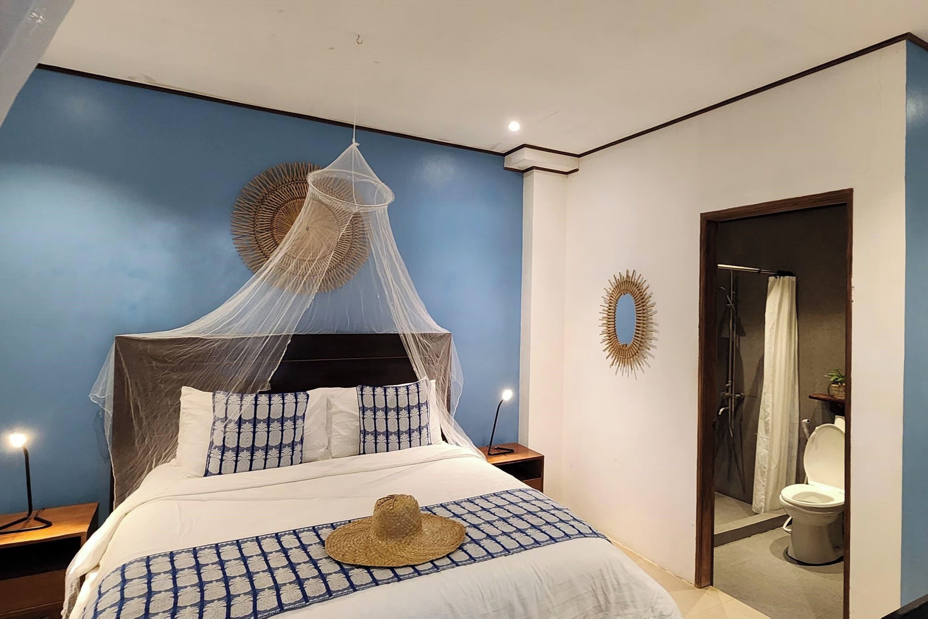 A bedroom with a bed, a nightstand, a bedside lamp, a decorative wall mirror, and a bathroom door.