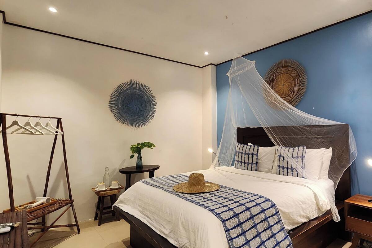 A bedroom with a bed under a mosquito net, a bedside table, a hanging clothes rack, and decorative wall art.