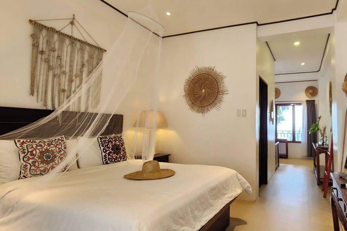 A bedroom with a bed, a hanging canopy, decorative pillows, a wall hanging, and a hallway leading to another room.