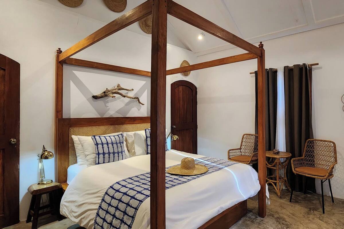 A four-poster bed with a wooden frame, a bedside table with a lamp, and two wicker chairs.