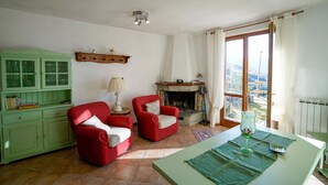 Un salon doté d’une cheminée, de deux fauteuils rouges, d’un buffet vert et offrant une vue sur l’extérieur grâce à une grande fenêtre.