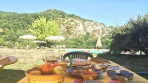 An outdoor dining setup with a table set for a meal, surrounded by chairs and umbrellas, with a mountainous backdrop. - Apartment in Marche near Urbino and Pool (Apecchio)