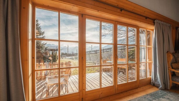 A room with large wooden-framed windows, a wooden deck outside, and a cozy interior with a rug and a chair. - Chalet in Kitzbühel near Ski Lifts (Kitzbühel)