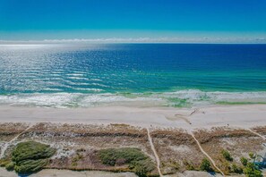 An aerial view of a beach with clear blue waters, sandy shore, and sparse vegetation.
