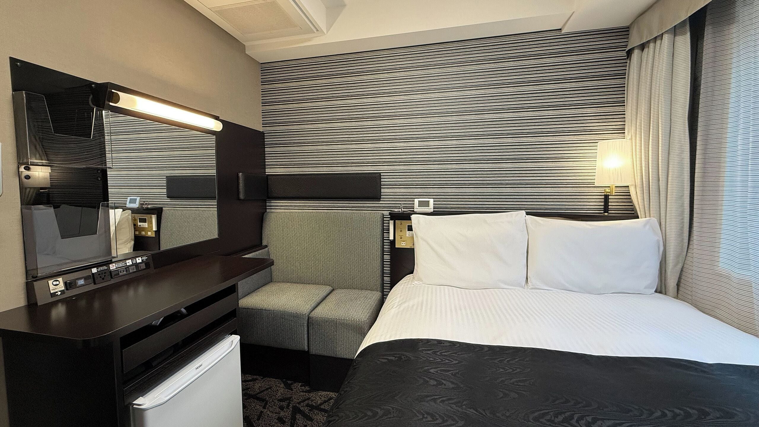 Ein Hotelzimmer mit Bett, Schreibtisch mit Spiegel, Stuhl, kleinem Kühlschrank und Wand-Fernseher.