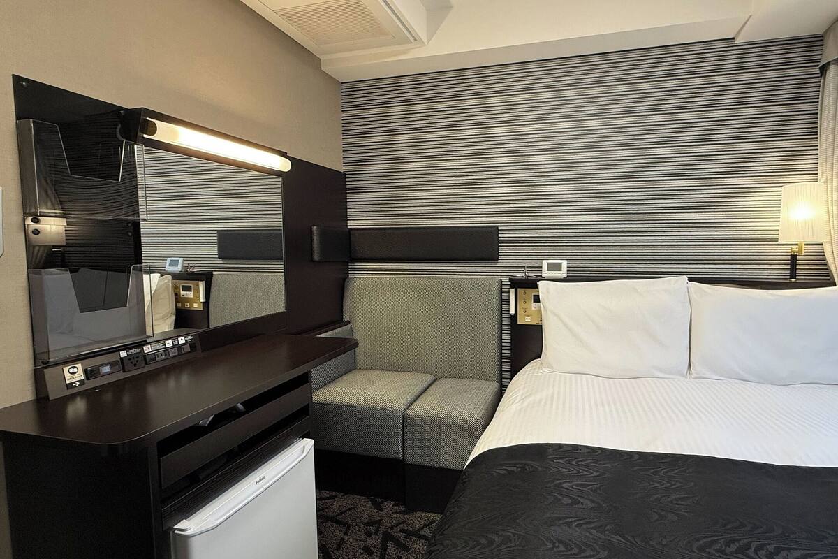 A hotel room with a bed, a desk with a mirror, a chair, a small refrigerator, and a wall-mounted TV.