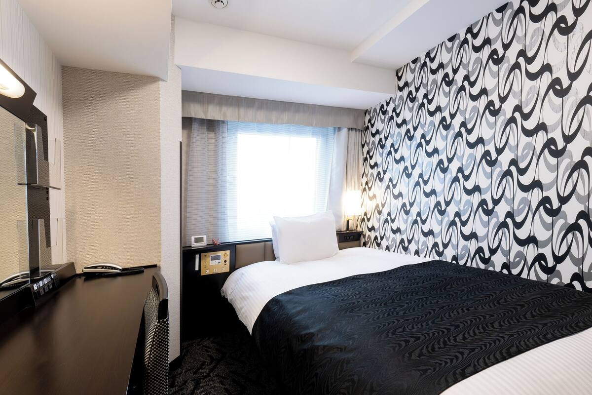 A hotel room with a bed, a desk, a chair, a nightstand, a lamp, and a patterned wall.