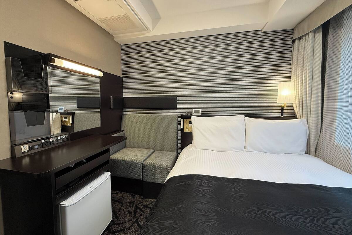 A hotel room with a bed, a desk with a refrigerator, a seating area with a chair, a large mirror, and a wall with horizontal stripes.