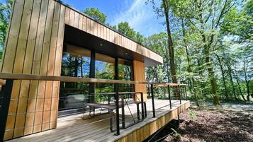 A modern wooden cabin with a deck, large windows, and a forest backdrop.