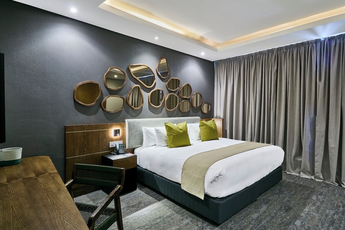 Una habitación de hotel moderna con una cama grande, una mesita de noche de madera, un escritorio y una silla. Hay varios espejos en la pared y un ventanal con cortinas.