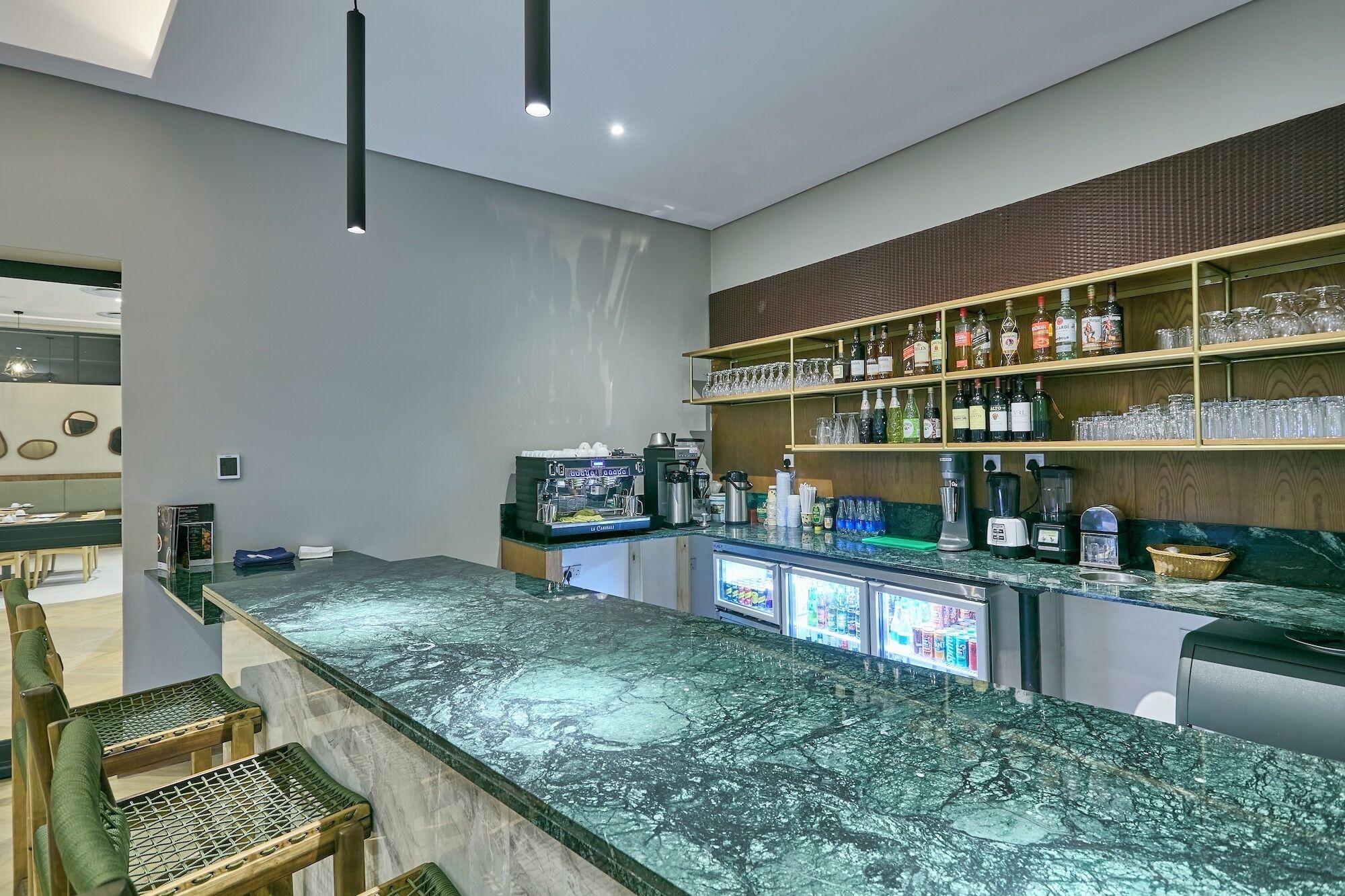 Un espace bar doté d’un comptoir en marbre, d’étagères remplies de bouteilles et de verres, et d’un coin salon avec des chaises.