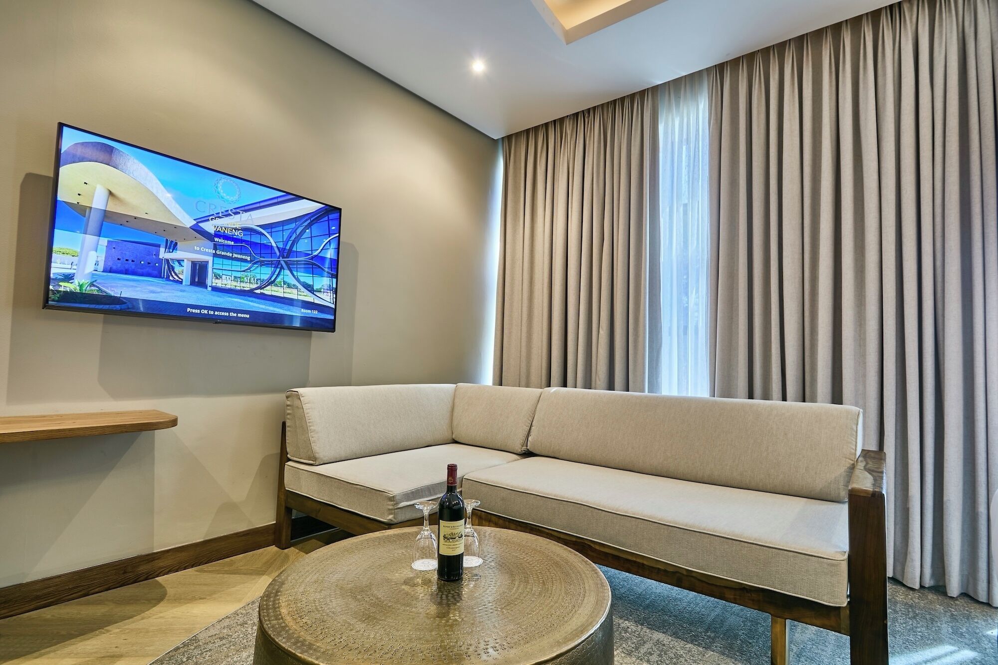 A modern living room with a beige sofa, a round coffee table with a bottle and glass, a flat-screen TV on the wall, and a large window with curtains.