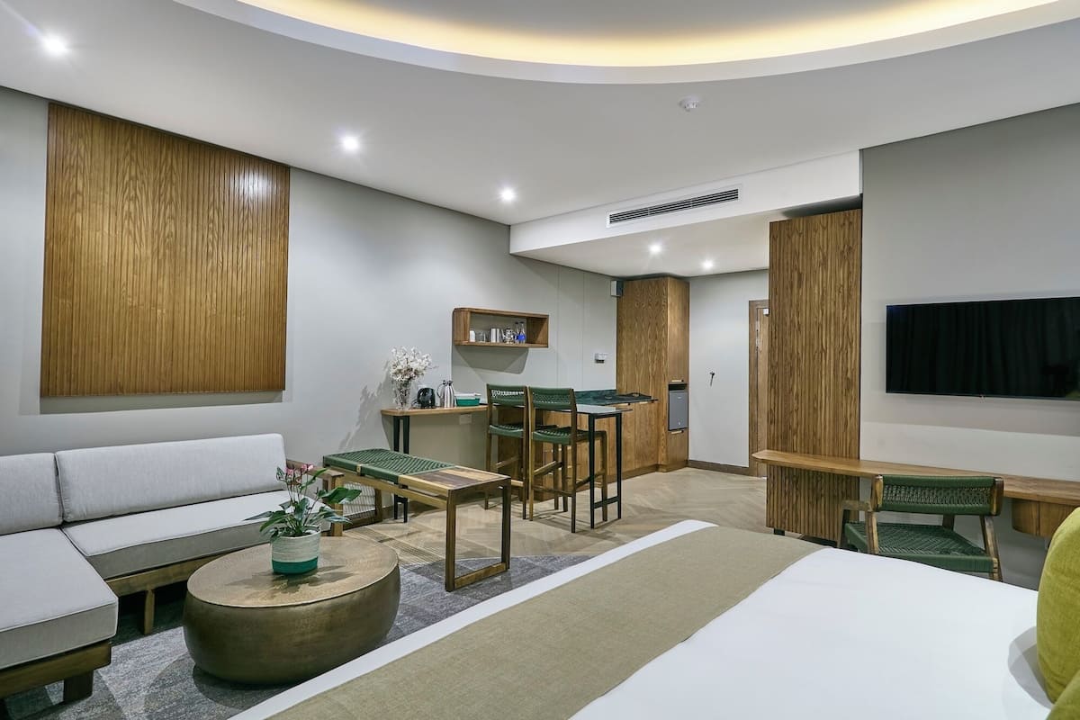Una habitación de hotel moderna con sofá, una mesa redonda con una planta, zona de comedor con mesa y sillas, televisor montado en la pared y paneles de madera en las paredes.