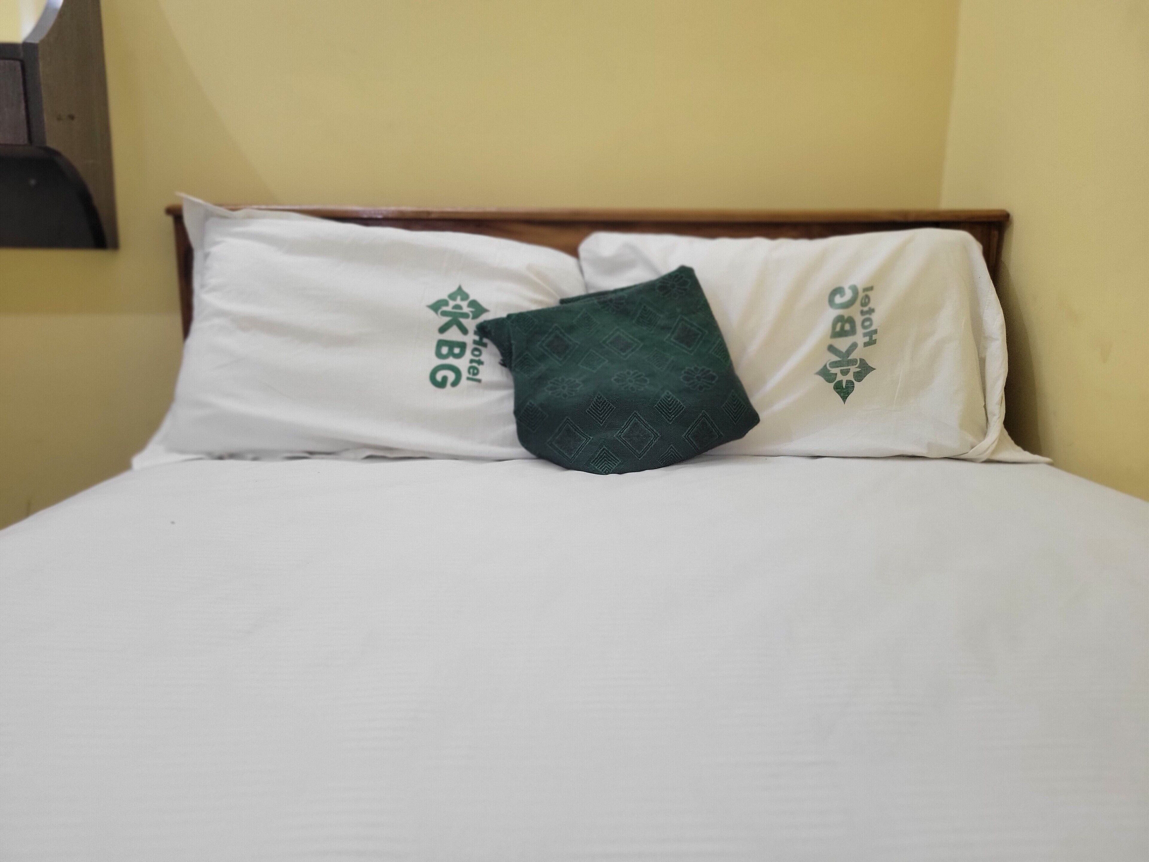 A bed with white bedding and pillows featuring the KBG Hotel logo.