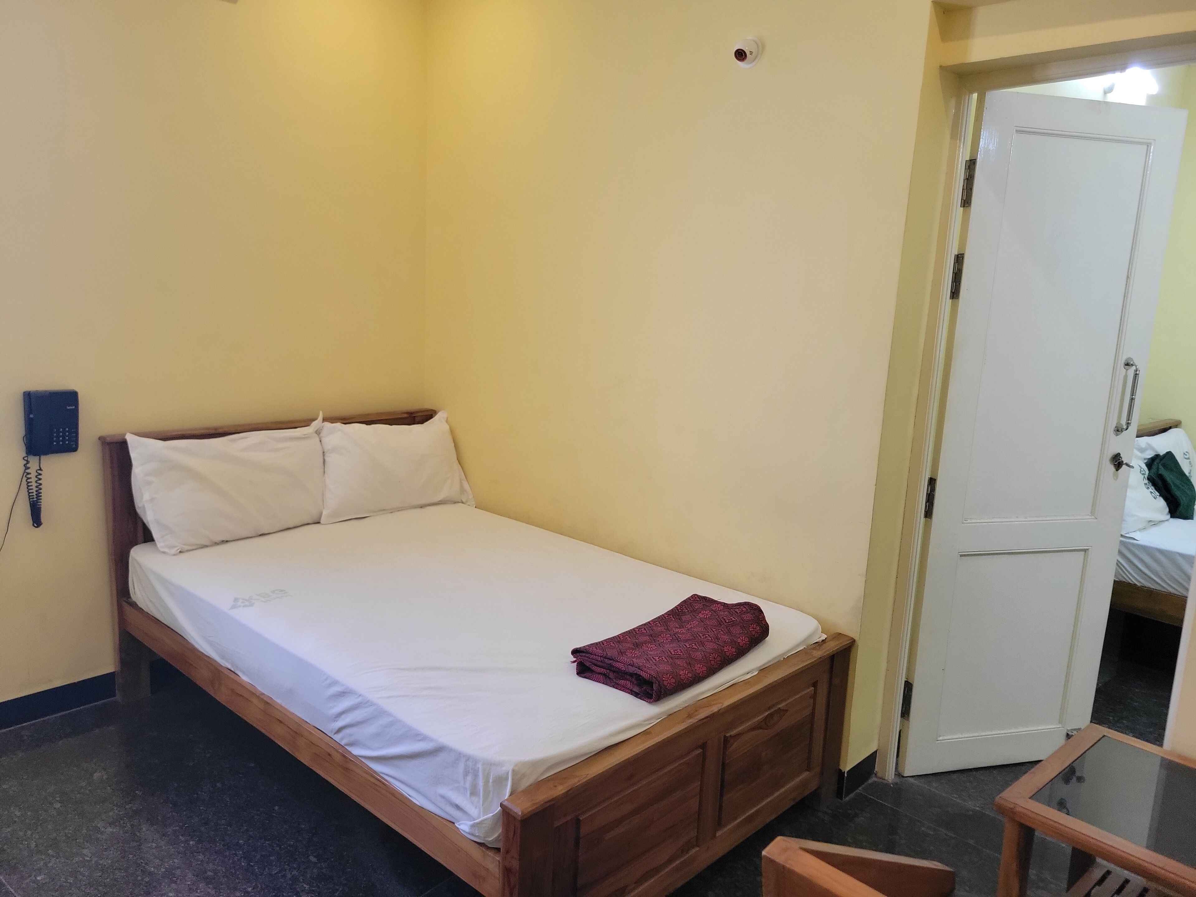 Uma cama de solteiro com estrutura de madeira, roupa de cama branca e um cobertor dobrado por cima. Um telefone está fixado na parede à esquerda da cama.