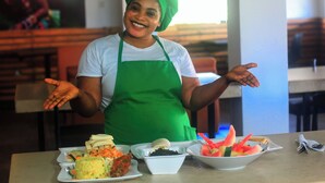 A person in a green apron and hat standing behind a table with various dishes. - Five Clover Hotel Monestery Road (Lekki)