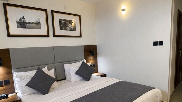 A modern hotel room with a large bed, two bedside tables, a nightstand, a wall-mounted light, and two framed pictures. - Five Clover Hotel Monestery Road (Lekki)